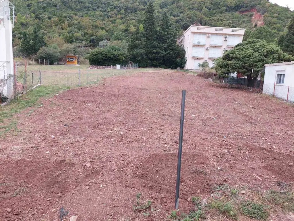 Cleared dirt lot with fence, buildings, and hillside backdrop