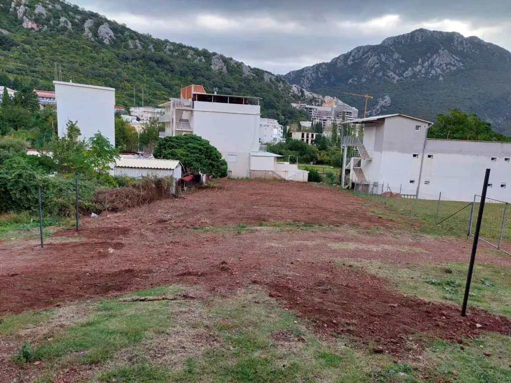 Cleared red-brown lot between white buildings and mountains