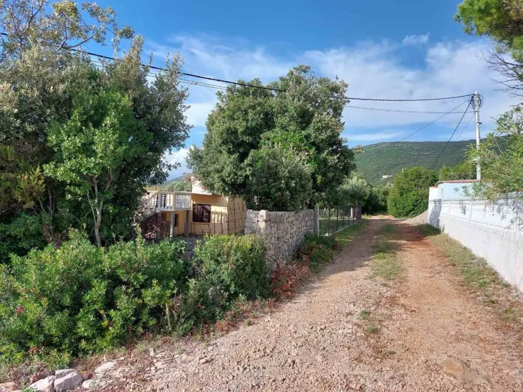 Dirt road beside stone wall and trees, with house visible