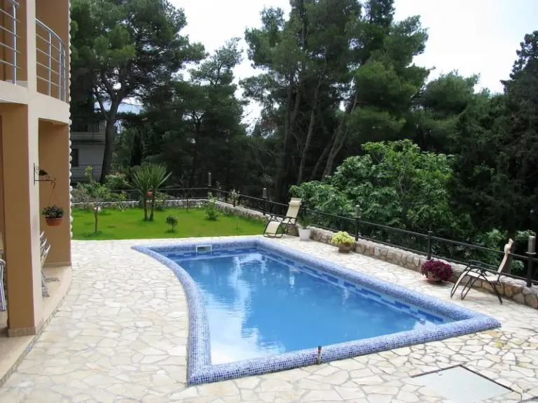 Curved blue-tiled swimming pool beside stone patio and trees