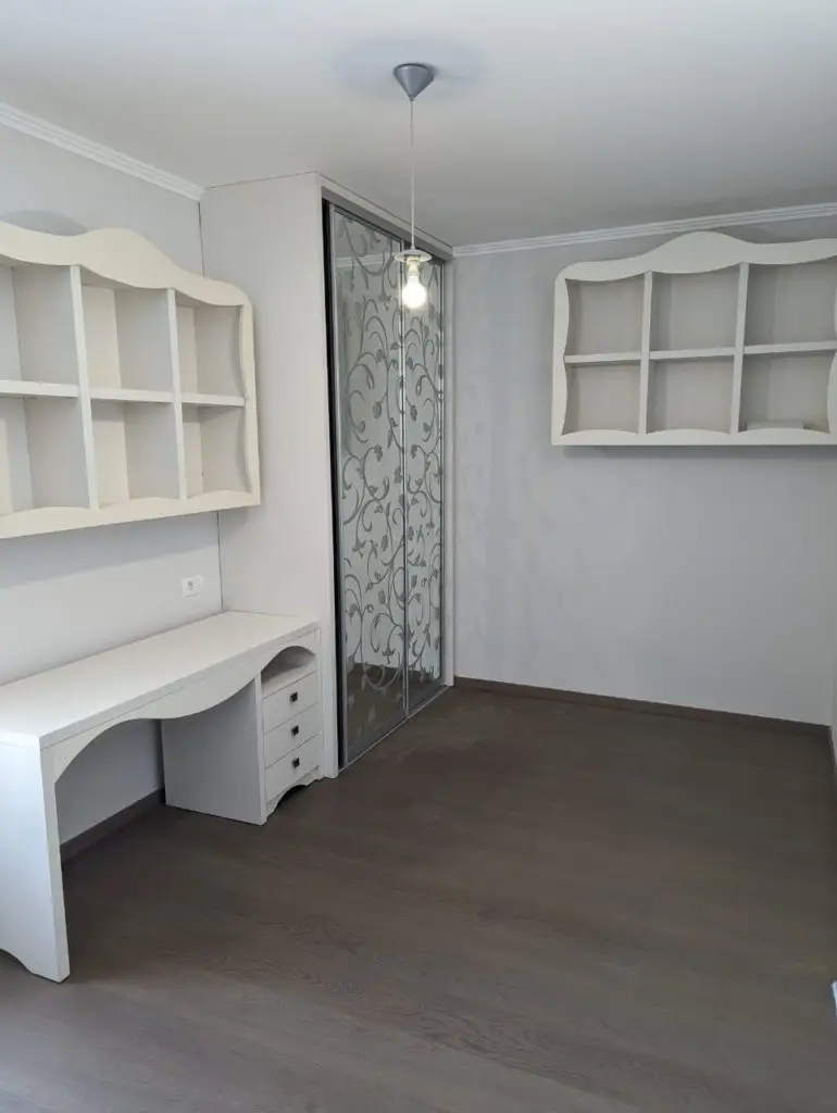 Small empty room with white desk, shelves, and wardrobe