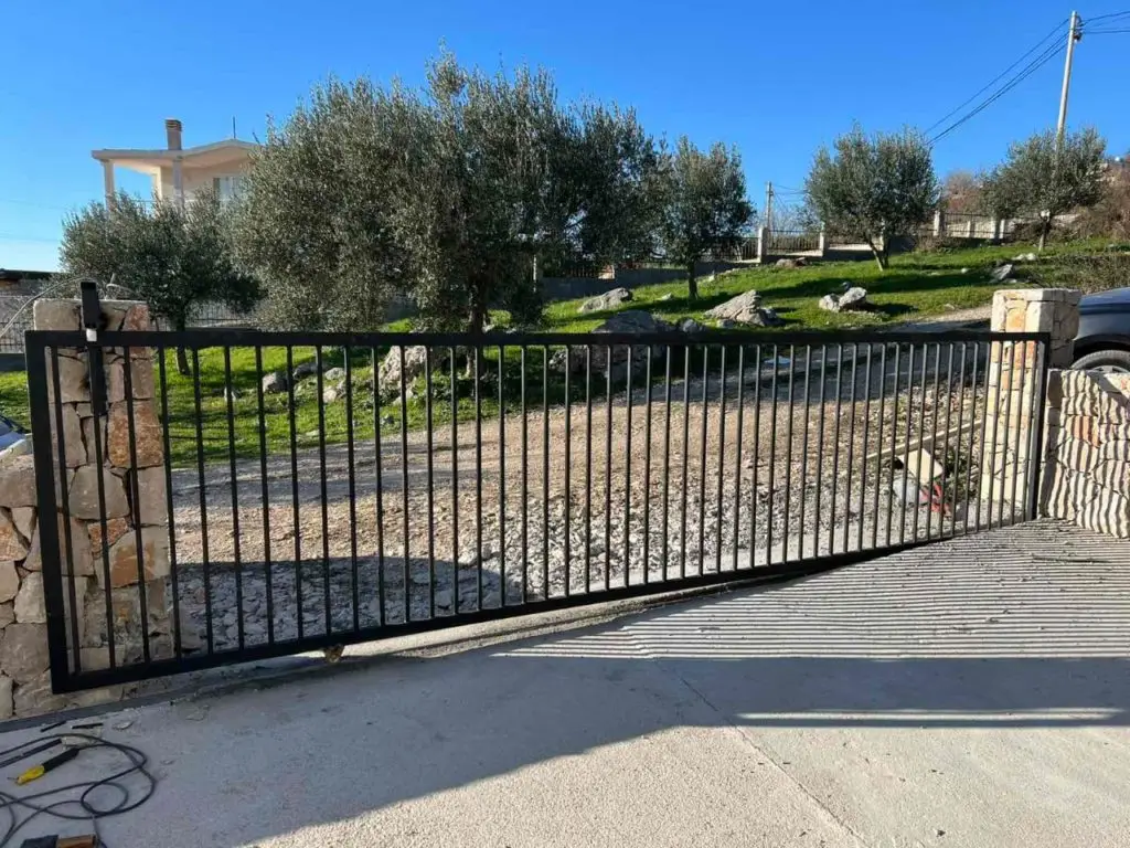 Black metal driveway gate with stone pillars and hillside behind
