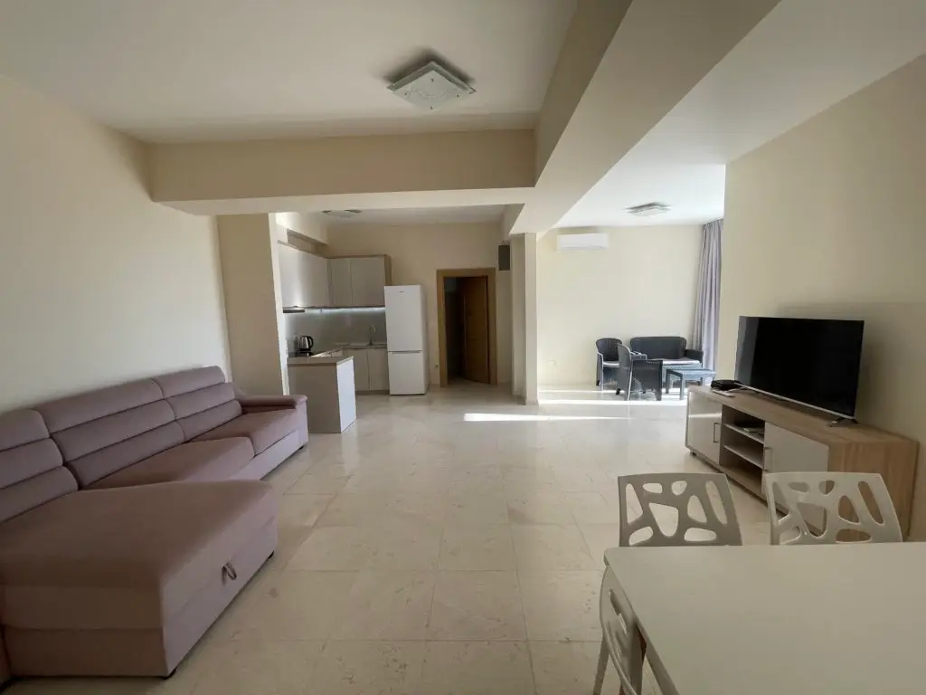 Open-plan living room with beige sofa, kitchen, TV, and dining table