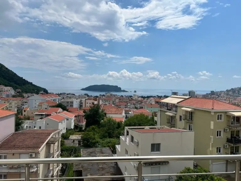 Balcony view over red-roofed buildings, sea, and island
