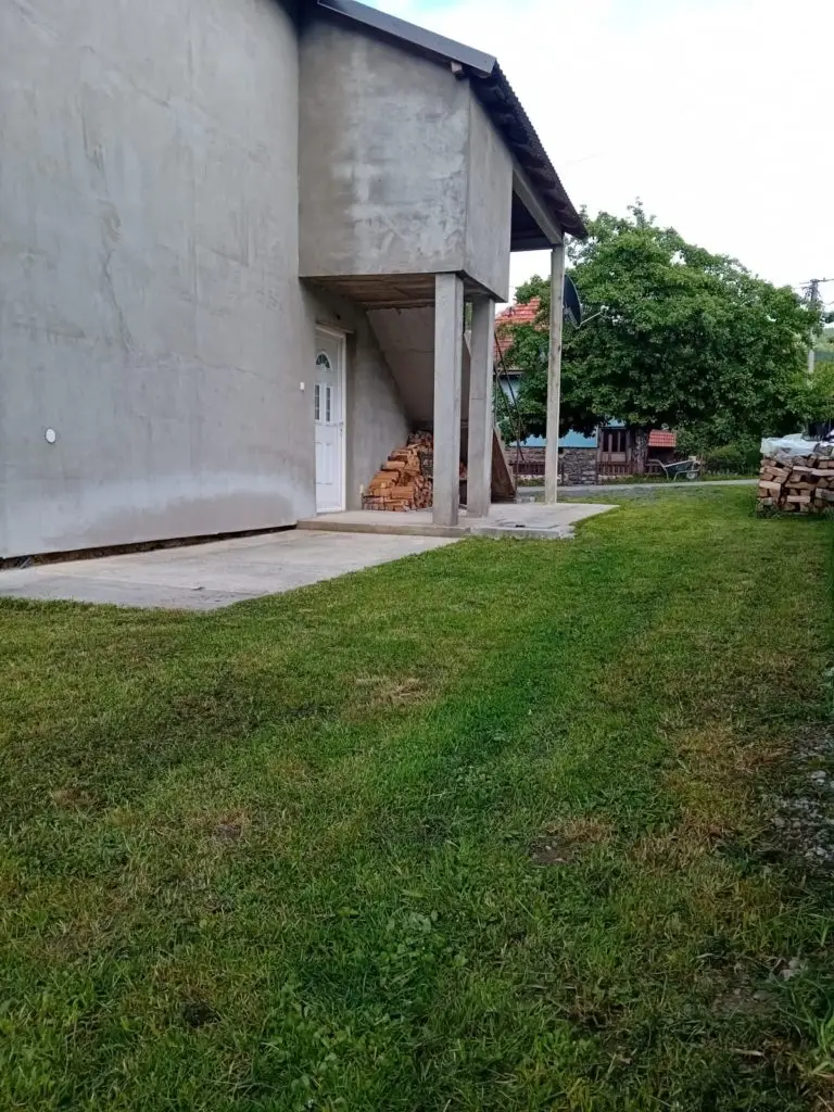 Concrete house exterior with porch, grass yard, and stacked firewood