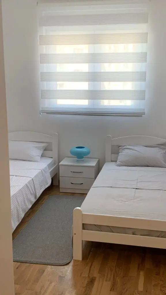 Two white single beds beside a small nightstand and window blinds