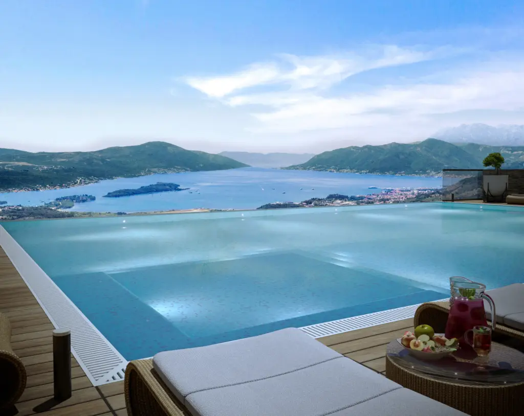 Infinity pool overlooking a bay and mountains, with lounge chairs and fruit on a table