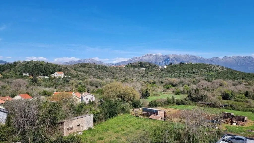 Rural landscape with houses, green hills, and mountains under blue sky