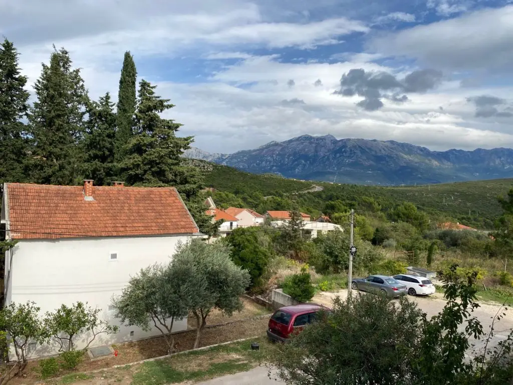 Rural house with red-tiled roof, trees, parked cars, mountains