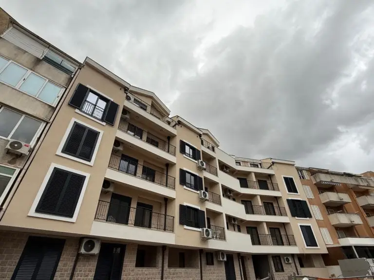 Beige apartment building with balconies and black shutters under cloudy sky