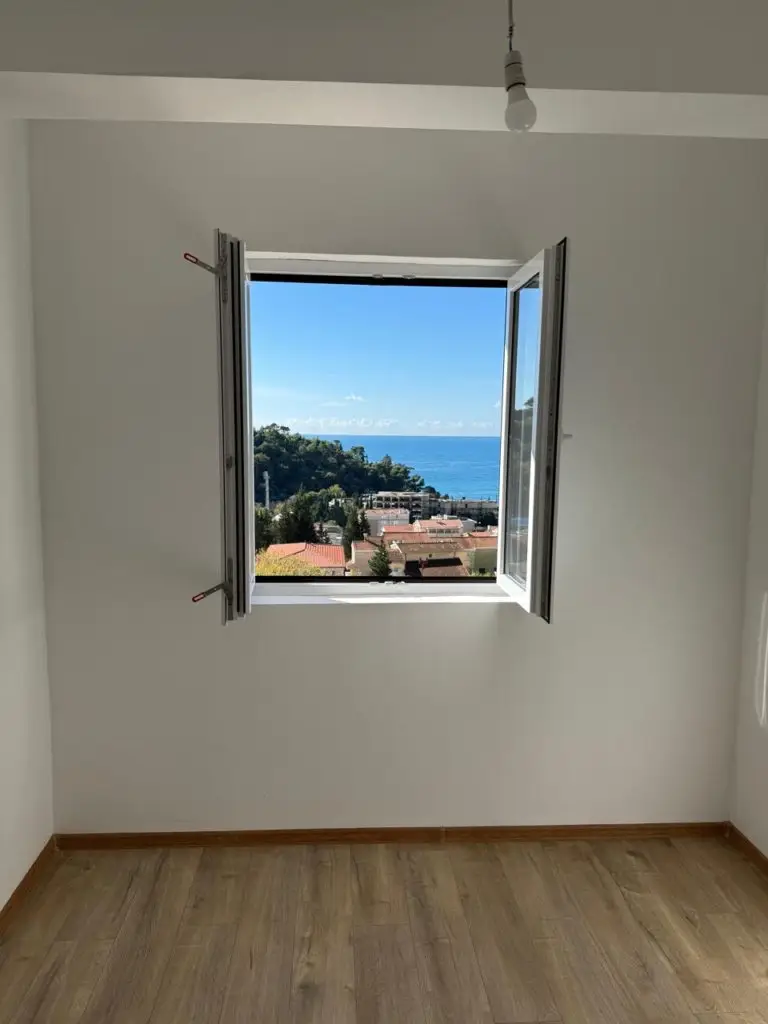 Empty room with open window overlooking sea and rooftops