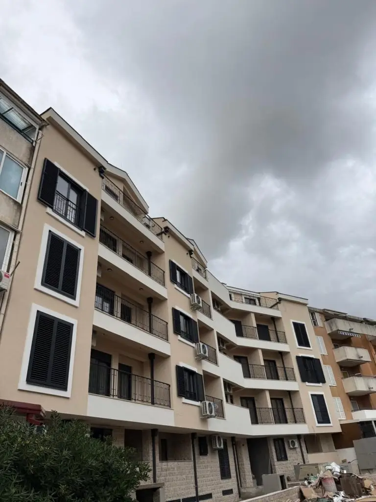 Beige apartment building with balconies and black shutters under cloudy sky