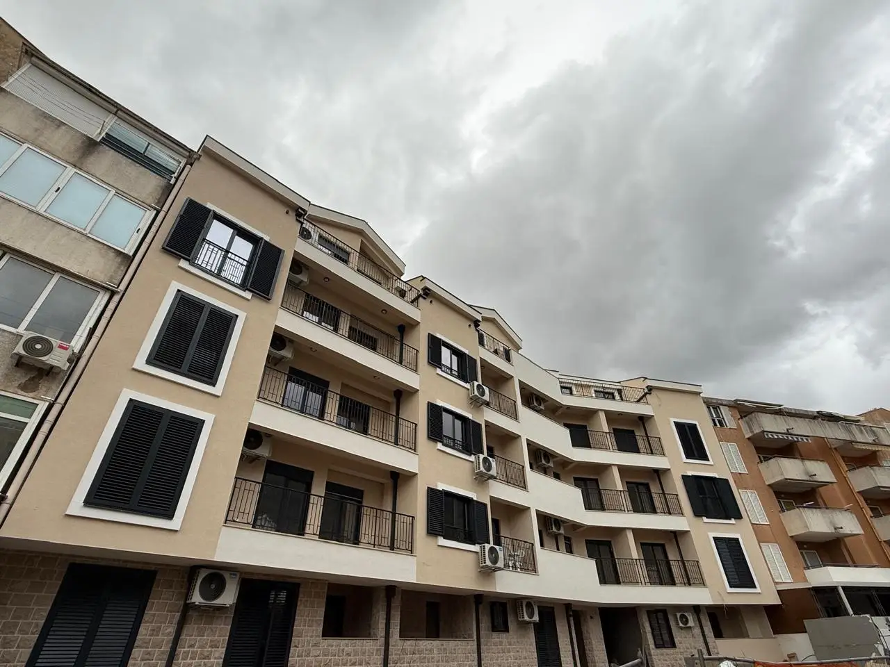 Multi-story apartment building with balconies and black shutters under cloudy sky