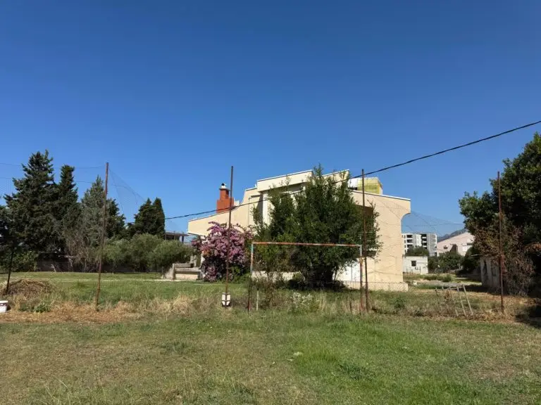 Cream-colored house behind grassy lot and trees under blue sky