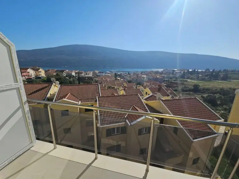 Balcony with glass railing overlooking rooftops, sea, and mountains